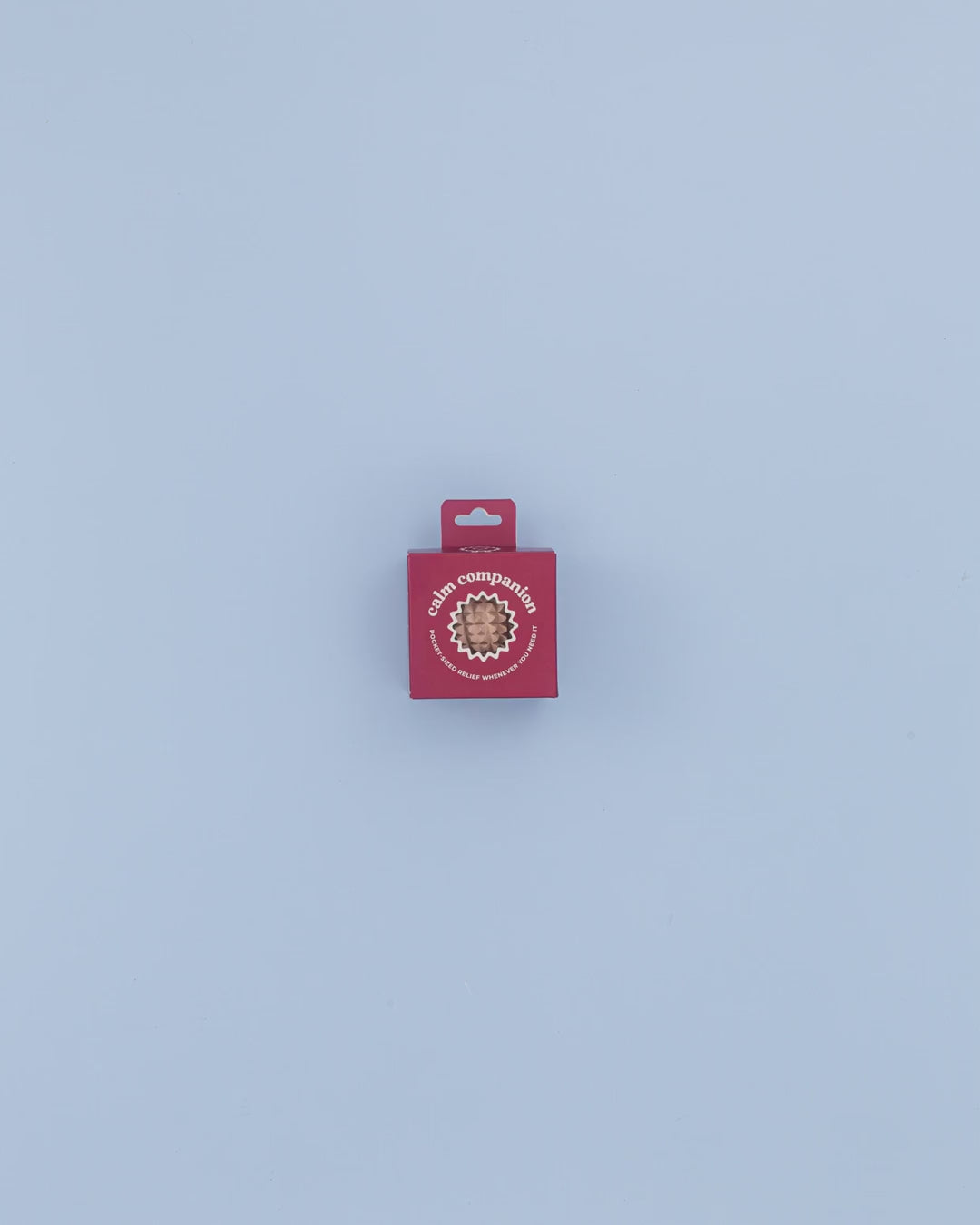 Small red box with a circular cutout revealing a textured stress relief ball inside, labeled 'Calm Companion™' for anxiety and stress relief, on a light blue background.